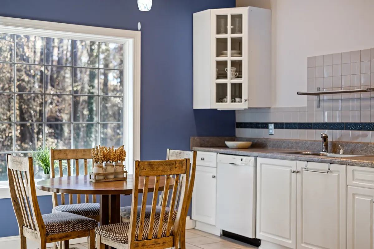 A bright kitchen with a round wooden dining table, white cabinets, and large window.